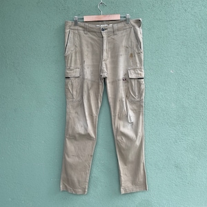 Puede incluir: Pantalones cargo color beige con múltiples bolsillos y cierre de botón. Los pantalones muestran signos de desgaste, incluyendo manchas y parches. Cuelgan de una percha metálica sobre un fondo turquesa.