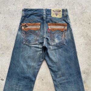 May include: Blue denim jeans with brown suede-like pocket accents and a red tag. The back pockets feature a distinctive stitched design. The Edwin brand label is visible at the waistband. The jeans have a classic, worn look.