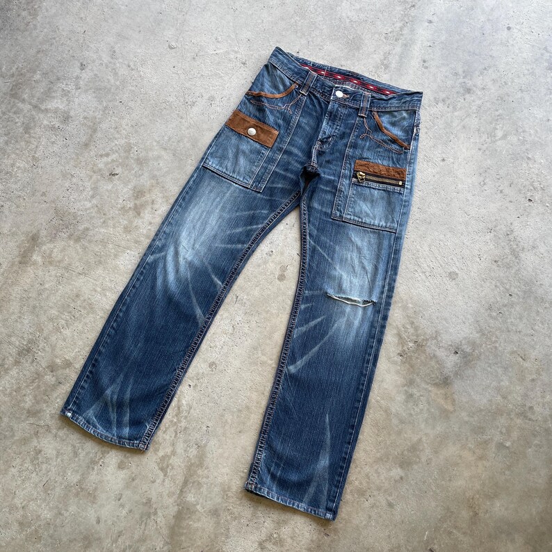 Puede incluir: Vaqueros azules de mezclilla con detalles de cuero marr&oacute;n en los bolsillos y un bolsillo con cremallera. Los vaqueros tienen un aspecto desgastado con un lavado descolorido y un peque&ntilde;o rasgu&ntilde;o en una rodilla. Cierre de bot&oacute;n y cremallera.