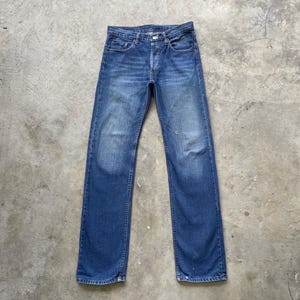 May include: Blue denim jeans with a button fly. The jeans have a classic five-pocket design and a straight leg fit. The denim has a faded wash with some wear and tear.
