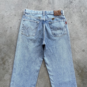 May include: Light blue denim jeans with a faded wash. The jeans feature classic five-pocket styling, a leather brand patch, and a red tag. The back pockets have a distinctive stitched design. The jeans are displayed on a gray surface.