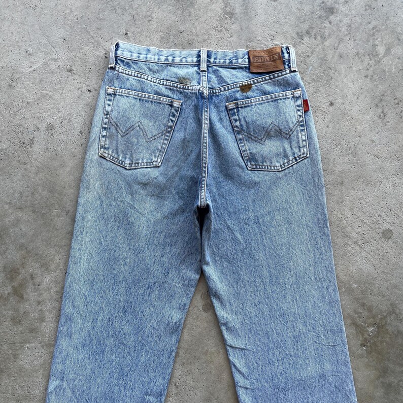 May include: Light blue denim jeans with a faded wash. The jeans feature classic five-pocket styling, a leather brand patch, and a red tag. The back pockets have a distinctive stitched design. The jeans are displayed on a gray surface.