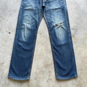 May include: Faded blue denim jeans with a distressed knee. The jeans have a classic five-pocket design and a straight leg fit. The denim has a worn appearance with lighter areas and fading throughout.