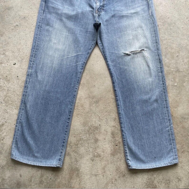 K&ouml;nnte beinhalten: Hellblaue Jeans mit einem abgenutzten, Used-Look. Die Jeans haben ein klassisches F&uuml;nf-Taschen-Design, einen Knopf- und Rei&szlig;verschluss und einen Riss am rechten Knie. Die Jeans liegen flach auf einer Betonoberfl&auml;che.