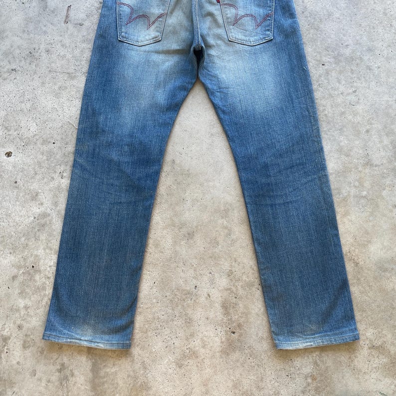 Puede incluir: Vaqueros de mezclilla azul descolorido con un dise&ntilde;o cl&aacute;sico de cinco bolsillos. Los vaqueros presentan un lavado claro con decoloraci&oacute;n sutil y marcas de desgaste. Los bolsillos traseros tienen detalles de costura rojos distintivos.