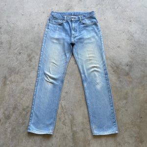 May include: Light blue denim jeans with a faded wash. The jeans have a classic five-pocket design, a button fly, and a straight leg. The jeans are laid flat on a concrete surface.