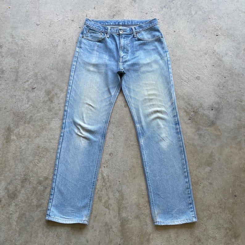 May include: Light blue denim jeans with a faded wash. The jeans have a classic five-pocket design, a button fly, and a straight leg. The jeans are laid flat on a concrete surface.