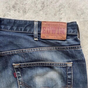 K&ouml;nnte beinhalten: Nahaufnahme der R&uuml;ckseite einer blauen Jeans. Ein braunes Lederetikett mit dem Markennamen "EDWIN" ist sichtbar, zusammen mit dem Text "GENUINE QUALITY JEANS FOR ALL THE PEOPLE OF THE WORLD BY EDWIN FOR CENTURIES TO COME". Die Jeans hat eine einzelne Ges&auml;&szlig;tasche.