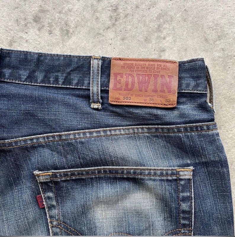 K&ouml;nnte beinhalten: Nahaufnahme der R&uuml;ckseite einer blauen Jeans. Ein braunes Lederetikett mit dem Markennamen "EDWIN" ist sichtbar, zusammen mit dem Text "GENUINE QUALITY JEANS FOR ALL THE PEOPLE OF THE WORLD BY EDWIN FOR CENTURIES TO COME". Die Jeans hat eine einzelne Ges&auml;&szlig;tasche.