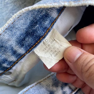 May include: Close-up of a light blue denim jeans tag. The tag reads "38" and "100% Cotton Made in U.S.A." The brand name "EDWIN" is also visible. The jeans are faded and show signs of wear.