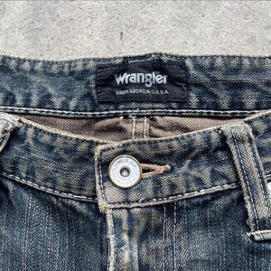 May include: Close-up of blue denim jeans with a button closure. The jeans feature a Wrangler label at the waistband, with the text "Wrangler GREENSBORO, N.C., U.S.A." visible. The jeans have a faded, worn appearance.