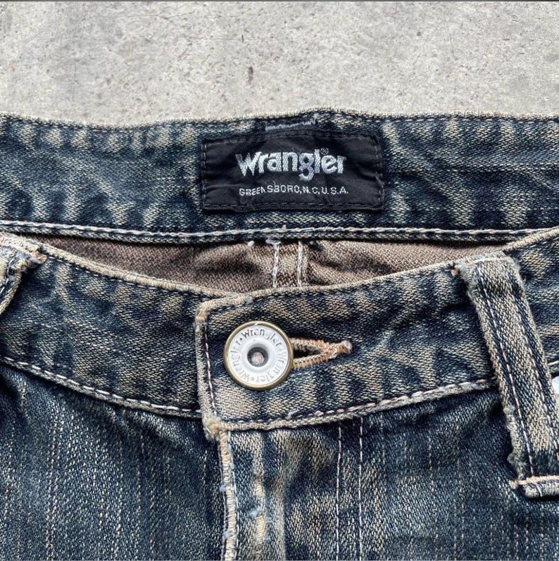 May include: Close-up of blue denim jeans with a button closure. The jeans feature a Wrangler label at the waistband, with the text "Wrangler GREENSBORO, N.C., U.S.A." visible. The jeans have a faded, worn appearance.