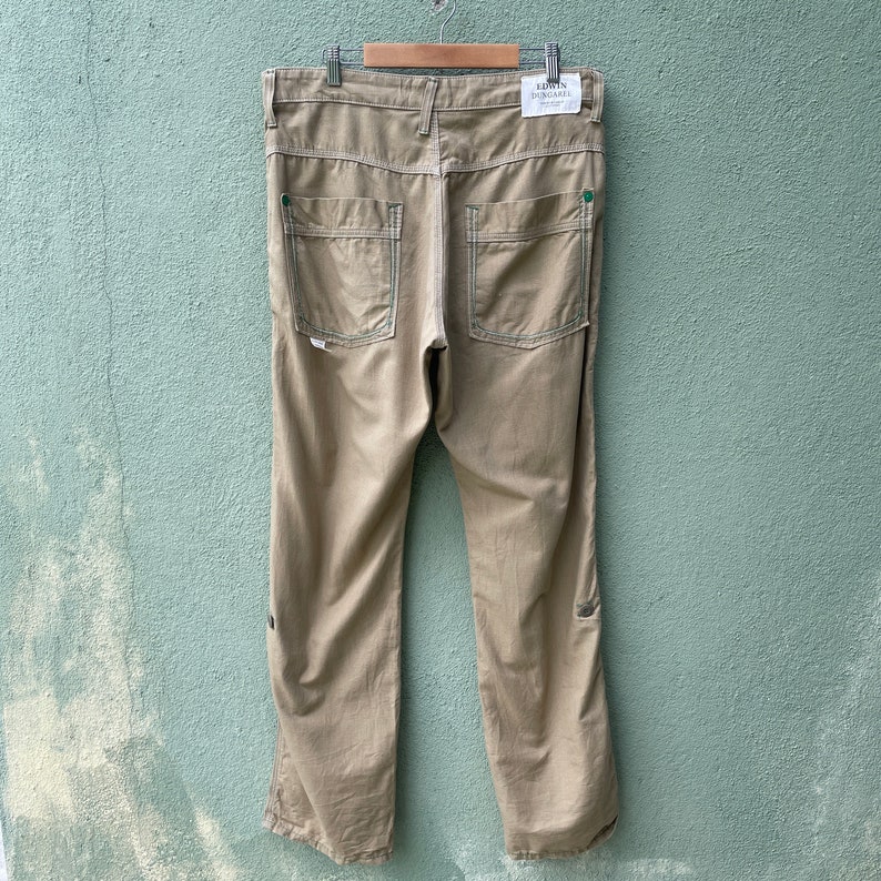 May include: Khaki-coloured trousers with green stitching and pocket accents. The trousers have two back pockets and a small tag on the back. The trousers are hanging on a metal hanger.