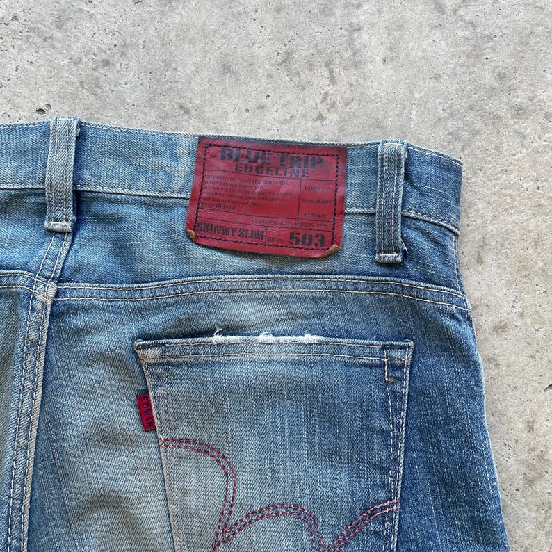 Puede incluir: Primer plano de la parte trasera de unos vaqueros azules. Los vaqueros presentan una etiqueta de cuero roja con el texto "BLUE TRIP EDGELINE" y "SKINNY SLIM 503". Las costuras rojas acent&uacute;an el bolsillo trasero y las costuras.