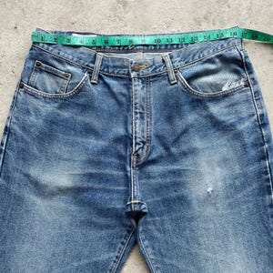 May include: Blue denim jeans with a classic five-pocket design. The jeans have a faded wash and a straight leg cut. A measuring tape is placed across the waist of the jeans, showing measurements in centimetres.