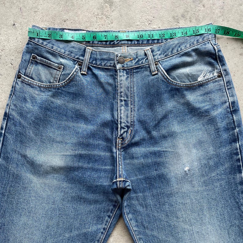 May include: Blue denim jeans with a classic five-pocket design. The jeans have a faded wash and a straight leg cut. A measuring tape is placed across the waist of the jeans, showing measurements in centimetres.