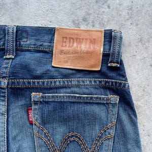 May include: Close-up of the rear of blue denim jeans. A brown leather patch with the brand name "EDWIN" and "Exclusive Vintage" is stitched on the waistband. The back pocket features the "EDWIN" brand tag. The jeans have a classic five-pocket design.