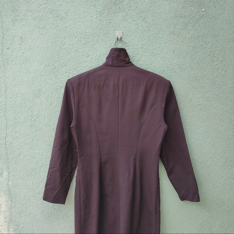 May include: A dark brown, long-sleeved dress with a high collar, hanging against a textured, light green wall. The dress is tailored with curved seams down the back, and is suspended from a metal hook.
