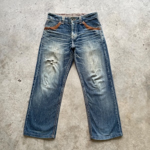May include: Blue denim jeans with a distressed look, featuring a faded wash and a ripped knee. The jeans have brown leather accents on the front pockets and a patterned waistband. The jeans are a classic five-pocket style.