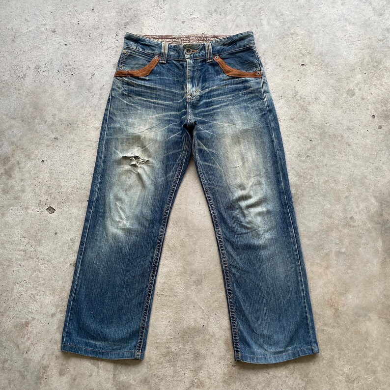 May include: Blue denim jeans with a distressed look, featuring a faded wash and a ripped knee. The jeans have brown leather accents on the front pockets and a patterned waistband. The jeans are a classic five-pocket style.