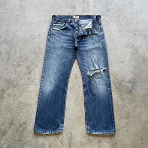 May include: Faded blue denim jeans with a button fly and distressed details. The jeans have a worn appearance with a large rip on the right knee and smaller rips on the pockets. The jeans are laid flat on a concrete surface.