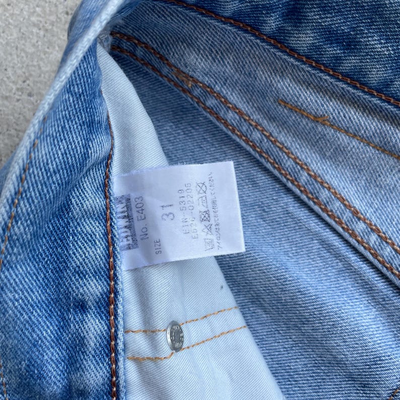May include: Close-up of a pair of blue denim jeans. A white tag is visible with the text "SIZE 31" and "No. E403". The jeans have visible stitching and a metal button. The tag also includes washing instructions.
