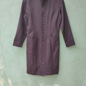 May include: A long, black dress with long sleeves and a button-up front. The dress features a mandarin collar, two front pockets, and a tailored fit. The dress is made of a smooth, solid fabric.