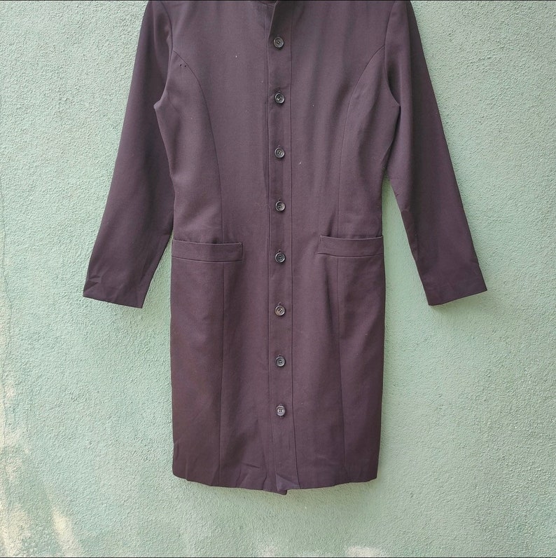 May include: A long, black dress with long sleeves and a button-up front. The dress features a mandarin collar, two front pockets, and a tailored fit. The dress is made of a smooth, solid fabric.