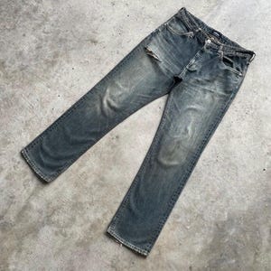 May include: Faded blue denim jeans with a distressed look. The jeans have a button closure, pockets, and a ripped section on the left leg. The jeans are laid flat on a concrete surface.