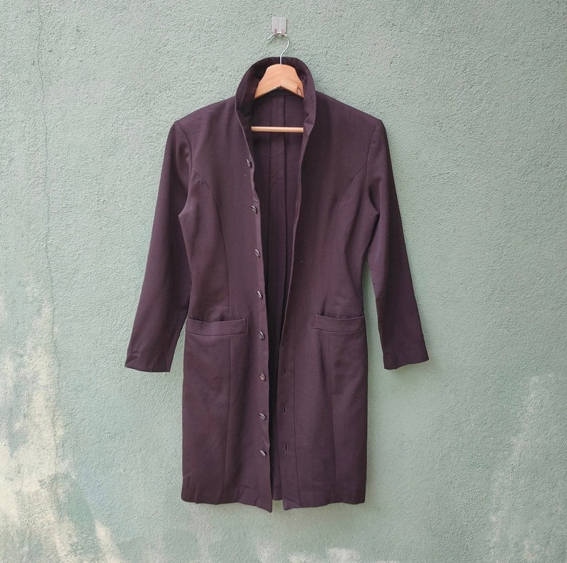 May include: A dark brown long coat with a mandarin collar and button-down front. The coat has two front pockets and long sleeves. It is hanging on a wooden hanger against a light green wall.