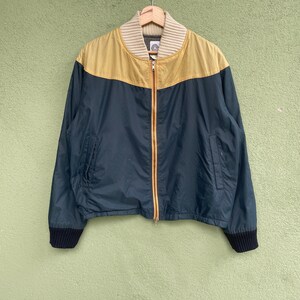 May include: A navy blue and tan bomber jacket with a ribbed collar and cuffs. The jacket has a full-length zipper and two front pockets. The top portion of the jacket is tan, while the bottom is navy blue. The jacket is hanging on a wooden hanger.