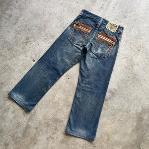 May include: Faded blue denim jeans with brown leather-like accents on the back pockets. The jeans have a classic five-pocket design and a small brand patch. The jeans are laid flat on a concrete surface.