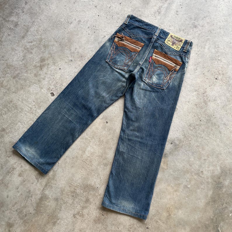 May include: Faded blue denim jeans with brown leather-like accents on the back pockets. The jeans have a classic five-pocket design and a small brand patch. The jeans are laid flat on a concrete surface.