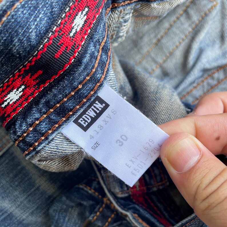 Puede incluir: Primer plano de unos vaqueros de mezclilla azules con un detalle interior estampado en rojo y blanco. Los vaqueros tienen una etiqueta que dice "EDWIN" y la talla 30. La mezclilla est&aacute; ligeramente descolorida.