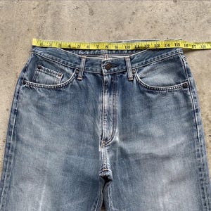 May include: Light blue denim jeans with a classic five-pocket design. The jeans are measured with a yellow measuring tape, showing a waist measurement of approximately 40.6 cm across. The jeans have a faded wash and a button and zipper closure.