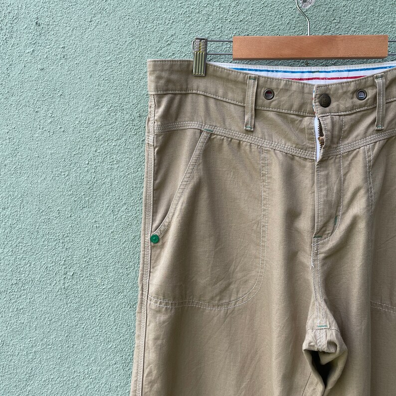 May include: Khaki-coloured trousers with prominent stitching and a button closure. The trousers feature front pockets and are suspended on a wooden hanger against a light green backdrop. The fabric appears to be hard-wearing.
