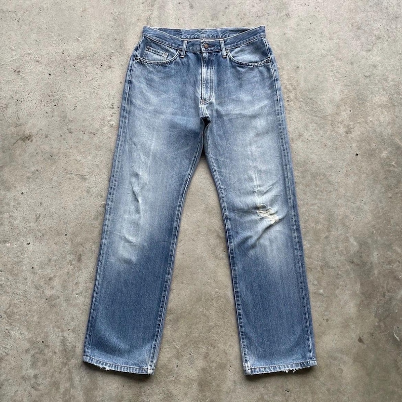 May include: Faded light blue denim jeans with distressed detailing. These jeans feature a classic five-pocket design and a straight leg cut. The denim has a worn look, including a ripped knee.