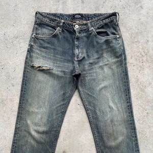 May include: Faded blue denim jeans with a distressed look, featuring a ripped knee. The jeans have a classic five-pocket design, a button and zipper closure, and a worn-in appearance. The jeans are a vintage style.