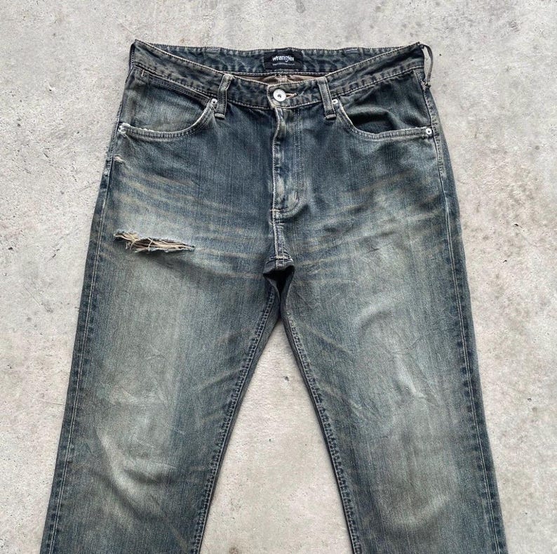 May include: Faded blue denim jeans with a distressed look, featuring a ripped knee. The jeans have a classic five-pocket design, a button and zipper closure, and a worn-in appearance. The jeans are a vintage style.