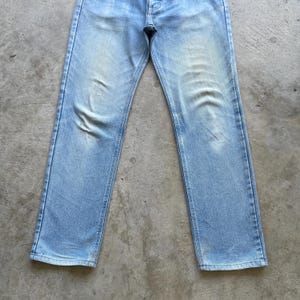 May include: Light blue denim jeans with a faded wash. The jeans have a classic five-pocket design, a button closure, and a straight leg fit. The denim shows signs of wear and fading, adding to the vintage aesthetic.