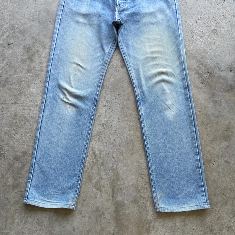 May include: Light blue denim jeans with a faded wash. The jeans have a classic five-pocket design, a button closure, and a straight leg fit. The denim shows signs of wear and fading, adding to the vintage aesthetic.