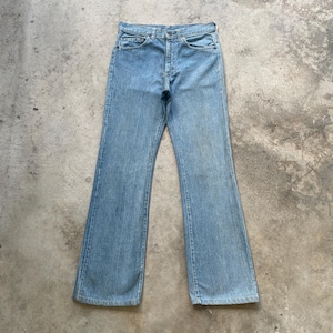 May include: Light blue denim jeans with a classic five-pocket design and a button-fly closure. The jeans have a flared leg opening and are made of a medium-wash denim material. The jeans are laid flat on a concrete surface.
