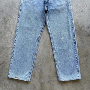 May include: Light blue denim jeans with a faded wash. The jeans have a classic five-pocket design, a button and zipper closure, and visible stitching along the seams. The jeans are slightly worn, with some discoloration.