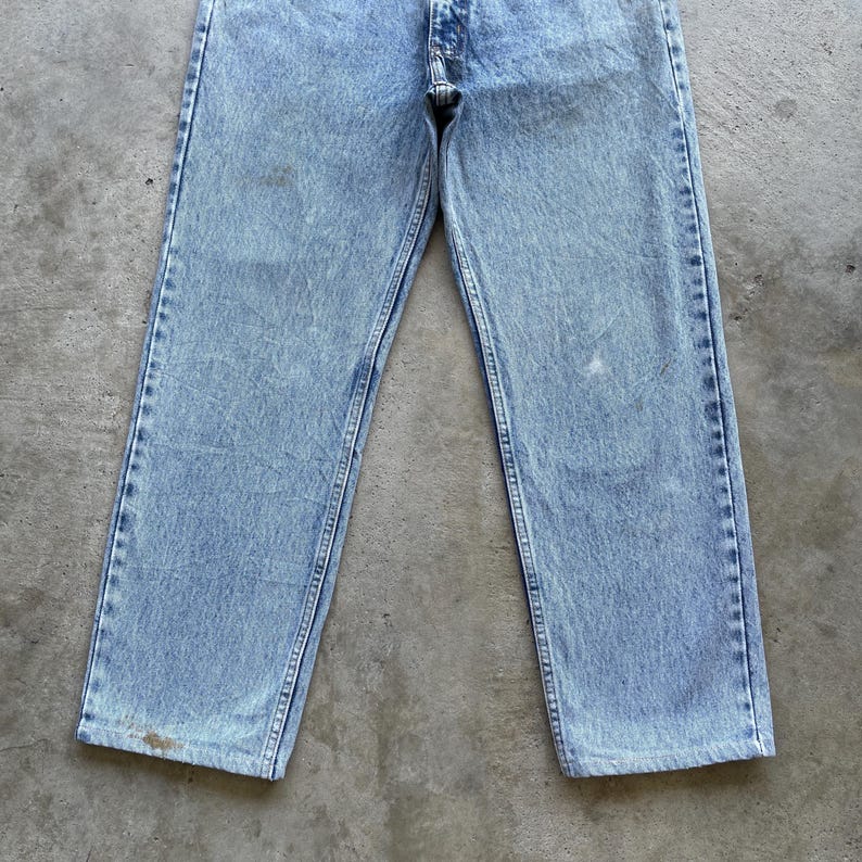 May include: Light blue denim jeans with a faded wash. The jeans have a classic five-pocket design, a button and zipper closure, and visible stitching along the seams. The jeans are slightly worn, with some discoloration.