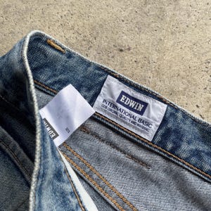 May include: Close-up of blue denim jeans. The jeans feature a white label with the brand name "EDWIN" and the text "INTERNATIONAL BASIC OUR ORIGINAL QUALITY AND WASHING". The jeans are stitched with yellow thread.