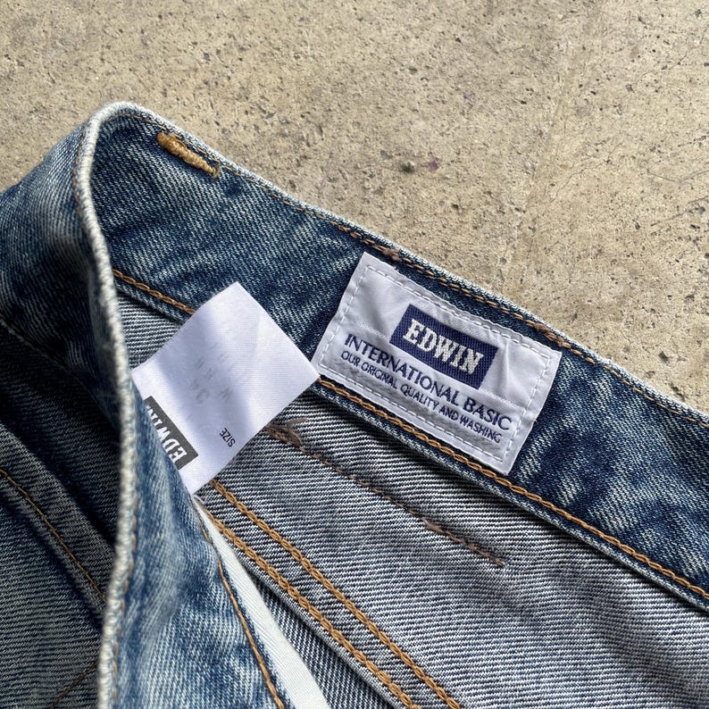 May include: Close-up of blue denim jeans. The jeans feature a white label with the brand name "EDWIN" and the text "INTERNATIONAL BASIC OUR ORIGINAL QUALITY AND WASHING". The jeans are stitched with yellow thread.