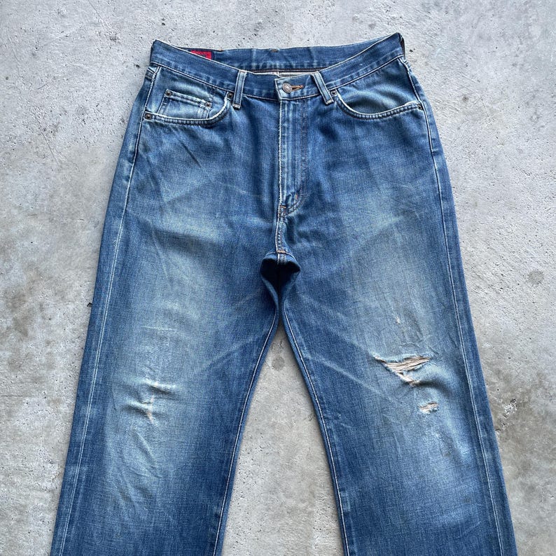 Pu&ograve; includere: Jeans in denim blu sbiadito con dettagli strappati, tra cui strappi e segni di usura. I jeans hanno un classico design a cinque tasche, una chiusura con bottone e cerniera e una vestibilit&agrave; dritta. Il denim ha un aspetto vintage e usurato.