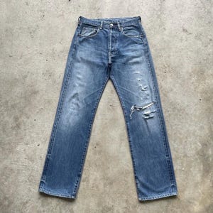May include: Faded blue denim jeans with a button fly and distressed details, including a large rip on the right knee. The jeans have a classic five-pocket design and a straight leg fit.