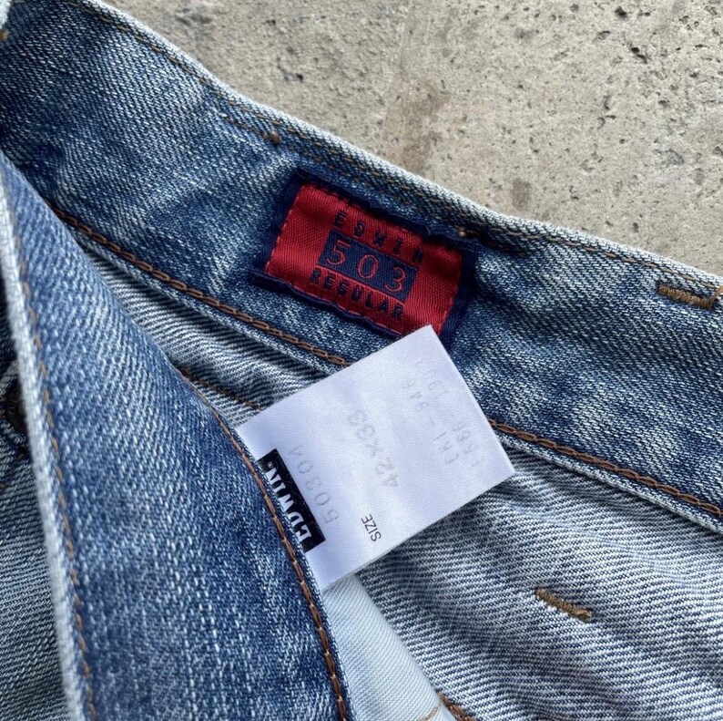 K&ouml;nnte beinhalten: Nahaufnahme einer hellblauen Jeans mit einem roten Edwin 503 Regular-Etikett. Ein wei&szlig;es Etikett zeigt die Gr&ouml;&szlig;e 42x33. Die Jeans hat ein klassisches F&uuml;nf-Taschen-Design und ein verwaschenes Aussehen.