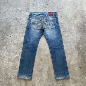 Puede incluir: Vaqueros de mezclilla azul descolorido con un dise&ntilde;o cl&aacute;sico de cinco bolsillos. Los vaqueros presentan un parche de cuero rojo en la parte trasera y costuras distintivas en los bolsillos traseros. Los vaqueros est&aacute;n colocados sobre una superficie de hormig&oacute;n.